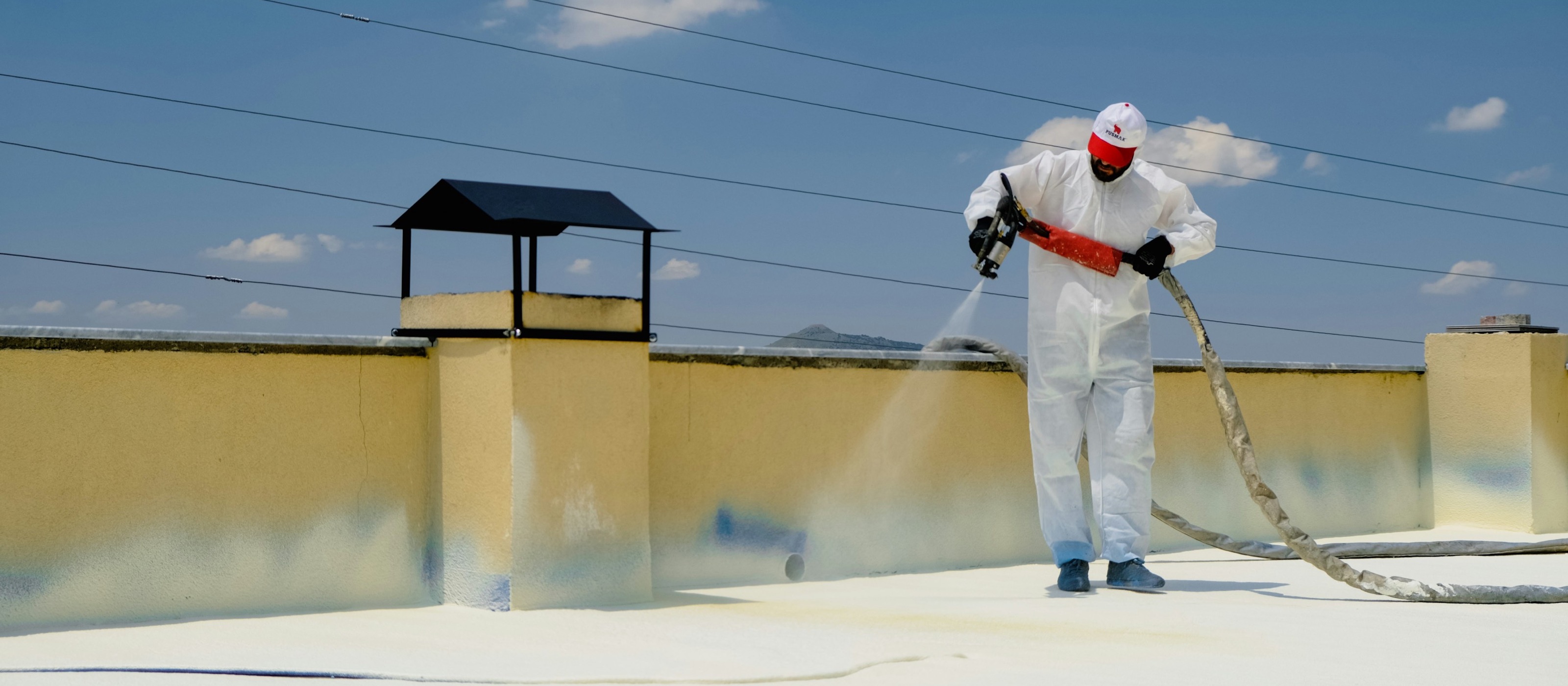 Applicator in white protective coverall spraying polyurethane foam across a flat roof deck under a clear sky